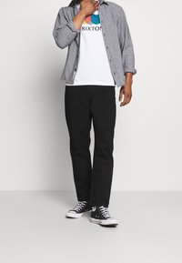 Pantaloni neri abbinati a una maglietta bianca con grafica e una camicia a quadri bianca e nera. Sneakers nere completano l'outfit.