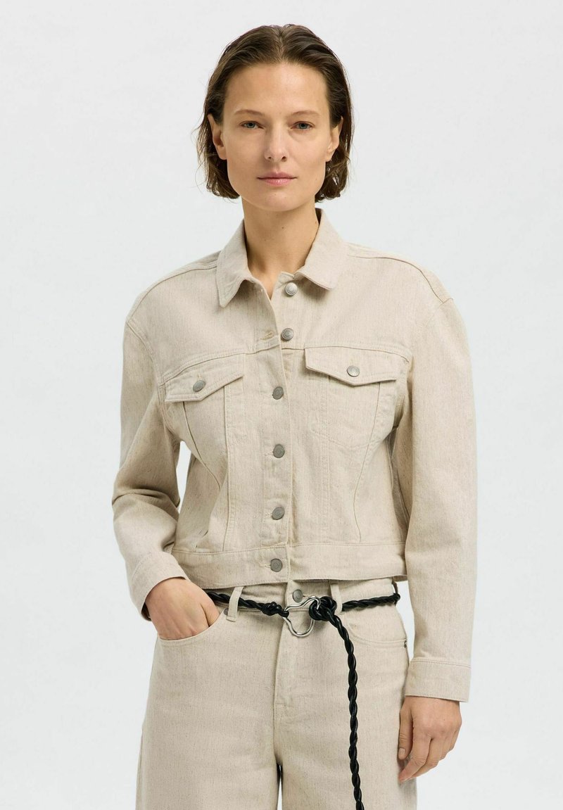 Chaqueta de mezclilla recortada beige con cierre de botones en la parte frontal, dos bolsillos en el pecho y una textura suave. Cuenta con un cinturón trenzado negro y costuras sutiles.