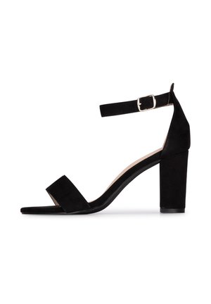 Sandalo alto in suede nero con cinturino alla caviglia, retro chiuso, fascia anteriore larga e tacco spesso. Texture liscia, dettagli minimi.