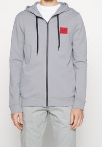 Hombre vistiendo una sudadera con cremallera de color gris claro, con cordones negros y un parche rojo de "HUGO", combinada con pantalones gris claro con bolsillos laterales.