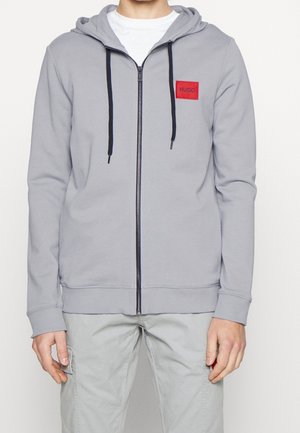 Man in een lichtgrijze hoodie met rits en zwarte trekkoorden, versierd met een rode 'HUGO'-patch, in combinatie met lichtgrijze broek met zijzakken.