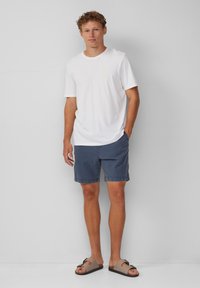Weißes Baumwoll-T-Shirt, kurze Ärmel, kleines Logo; kombiniert mit marineblauen Shorts, lockere Passform, gesäumte Kanten und hellbraunen Sandalen.