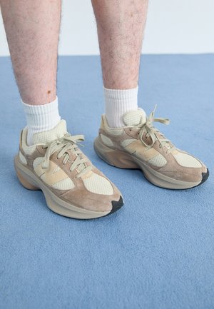 Beige and cream athletic shoes with mesh and suede materials, featuring thick soles and laces, worn with white crew socks on a blue surface.
