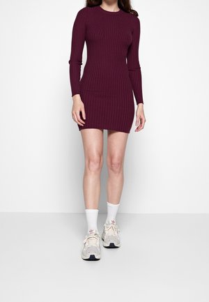 Robe pull de couleur bourgogne profond, texture côtes, manches longues, silhouette ajustée, assortie avec des chaussettes blanches et des baskets légères.