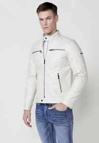 Chaqueta de cuero blanca con cuello alto, múltiples bolsillos con cremallera y hombros texturizados. Combinada con vaqueros azules. Diseño limpio y ajuste moderno.