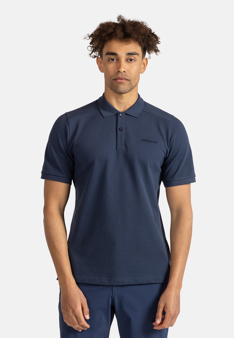 Námořnická modrá polo tričko vyrobené z hladké tkaniny, které se vyznačuje klasickým límečkem, dvěma knoflíky, krátkými rukávy a logem na levé hrudi.