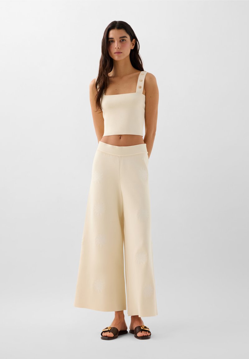 Cremfarbener Crop-Top mit quadratischem Ausschnitt und Knopf-Details, kombiniert mit weiten Hosen, die strukturierte Muster und ein weiches Strickfinish aufweisen.