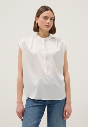 Donna con camicetta bianca senza maniche con colletto alla coreana e jeans blu, in piedi davanti a uno sfondo chiaro uniforme.