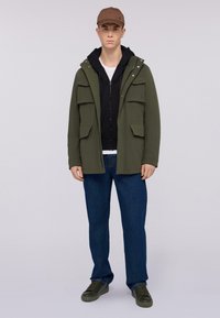 Zelený parka v olivové barvě s několika předními kapsami, navrstvená nad černou mikinou. V kombinaci s modrými džíny a tmavozelenými teniskami. Hnědý klobouk na hlavě.