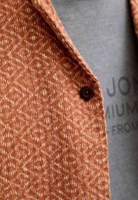 Gros plan sur une veste orange à motif géométrique avec un bouton foncé sur une chemise grise partiellement ornée de texte noir.