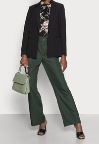 Blazer nero sopra una maglietta floreale, abbinato a pantaloni larghi verdi e sandali neri con tacco. Tiene in mano una borsa verde con manico testurizzato.
