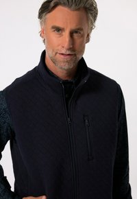 Mörkblå quiltad väst med hög krage, som har två dragkedjefickor och ett texturerat mönster. Bärs över en mönstrad mörk skjorta.