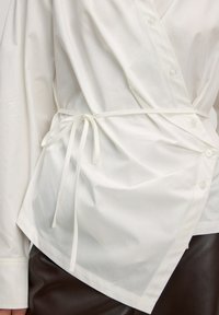 Witte wikkelshirt met een diagonaal ontwerp, lange mouwen, knoopdetails en een geknoopte taille-accent. Gladde stof met een lichte glans.