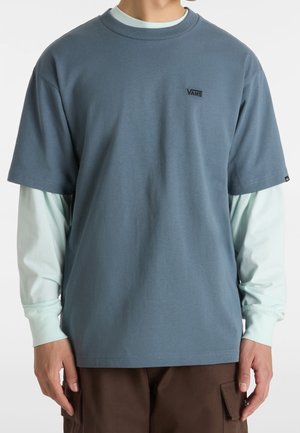 T-shirt bleu clair à manches courtes avec encolure ronde, arborant un petit logo noir sur la poitrine. Porté par-dessus une chemise à manches longues vert clair.