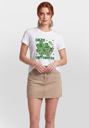 Vrouw met rood haar die een wit T-shirt draagt met een groene vierbladige klaver en een afbeelding van een ijskoud drankje en een beige rok, staand tegen een effen achtergrond.