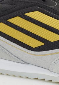 Schwarzer Sportschuh mit goldgestreiften Akzenten, strukturiertem Stoffobermaterial und hellgrauen Wildlederüberzügen. Das Markenzeichen "Messi" ist geprägt.