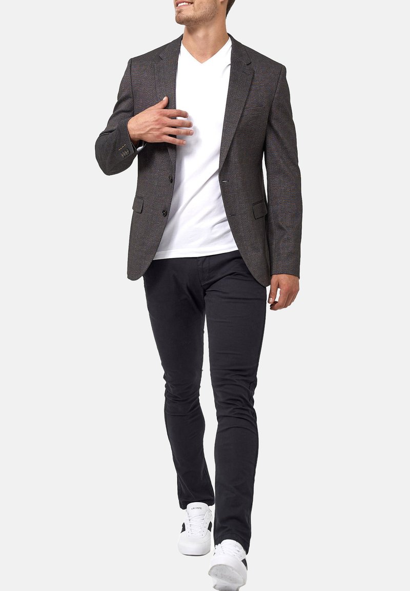 Blazer de estampado oscuro, camiseta blanca de cuello en V, pantalones slim-fit negros, zapatillas blancas. El blazer cuenta con dos botones frontales y bolsillos laterales.