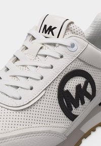 Λευκό δερμάτινο αθλητικό παπούτσι με διάτρητο σχέδιο, λευκά κορδόνια και μαύρο λογότυπο "MK" στο πλάι και στη γλώσσα, που δείχνει λεπτομερείς ραφές και υφή.