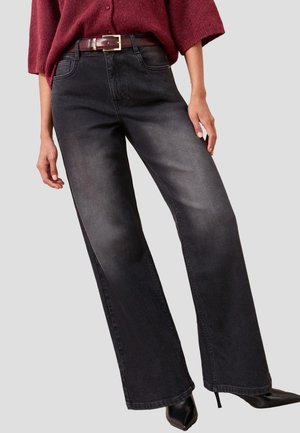 Femme portant un jean noir délavé à jambes larges, un pull bordeaux, une ceinture marron et des talons noirs pointus, debout devant un fond uni.