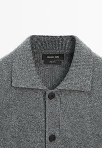 Cardigan in maglia di lana grigia con colletto piegato e due bottoni neri sul davanti, etichetta con dettagli sul marchio e sul materiale.