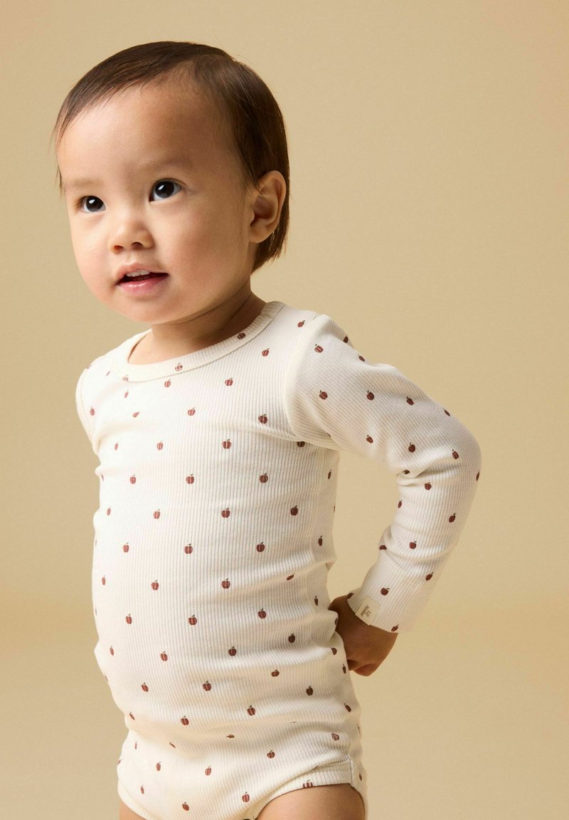 Tout-petit portant un body blanc à manches longues avec de petits motifs marron, debout les mains derrière le dos sur un fond beige.