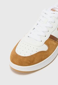 Lacoste sneaker fehér bőr felsőrészével, bézs nubuk részleteivel, perforált díszítéssel, lapos fehér fűzőkkel és gumitalppal rendelkezik.