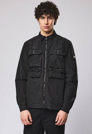 Młody mężczyzna z kręconymi włosami, ubrany w czarną kurtkę zapinaną na zamek z wieloma przednimi kieszeniami, stojący na tle jednolitego szarego tła.