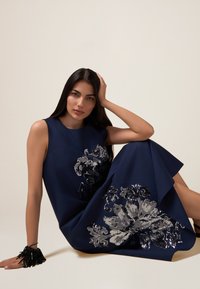 Marineblaues ärmelloses Kleid mit silberner Pailletten-Blumenstickerei, A-Linien-Form, strukturierter Stoff und asymmetrischem Saum. Schwarzes Quastenarmband.