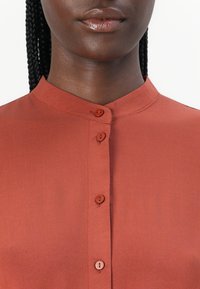 Blouse couleur rouille avec un col mandarin, comportant cinq boutons assortis et un tissu lisse, légèrement brillant.