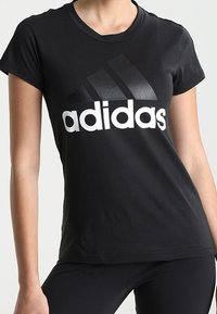 Černé bavlněné tričko s krátkým rukávem, na kterém je velké bílé logo "adidas" a tři černé diagonální pruhy na přední straně.