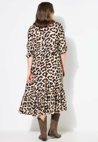 Leopardenmuster-Kleid mit einem gestuften Rüschensaum, aus einem weichen Stoff. Verfügt über Dreiviertelärmel und eine lockere Passform. Braune Stiefel sind sichtbar.