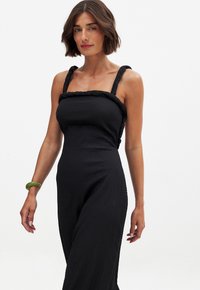 Svart jumpsuit med en texturerad tyg, flätade remmar och en åtsittande livdel som övergår till vida ben. Accenterad av ett grönt armband.