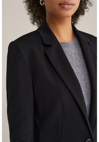 Crni sako krojenog dizajna s oštrim reverima i jednobotonim zatvaranjem, složen preko teksturiranog sivog džempera. Glatka, strukturirana tkanina.