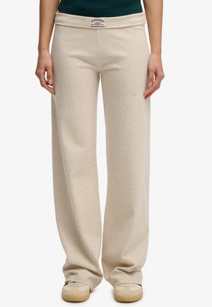 Beige wijde broek met Superdry-logo op de tailleband, gedragen met beige sneakers en een donkergroene top op een witte achtergrond.