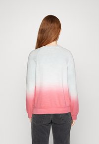 Persoon met lange rode haren die een sweatshirt met een wit-roze verloop en zwarte jeans draagt, gezien van achteren tegen een effen achtergrond.
