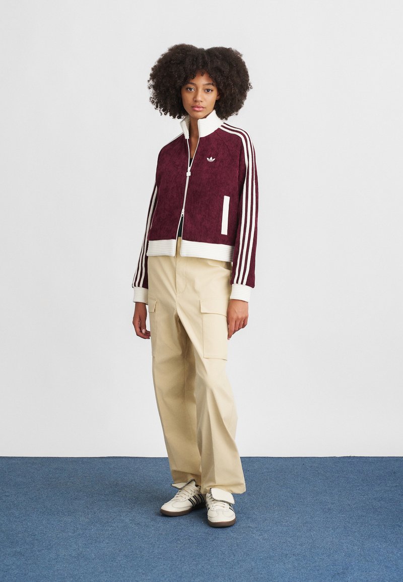 Bordeaux cropped jack met witte strepen, ritssluiting aan de voorkant, geribbelde manchetten en een geborduurd logo. Gepaard met beige cargobroek en witte sneakers.
