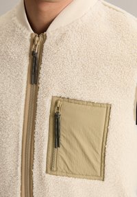 Crème en beige mouwloze vest met een zachte, gestructureerde fleece buitenkant, een ritssluiting aan de voorkant en een opgestikte zak met een ritssluiting.