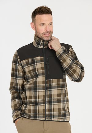 Man die een geruite flanellen jas met hoge kraag en ritssluiting aanpast, met een zwart borstpaneel en ritssluitingzakken, draagt khakibroek.