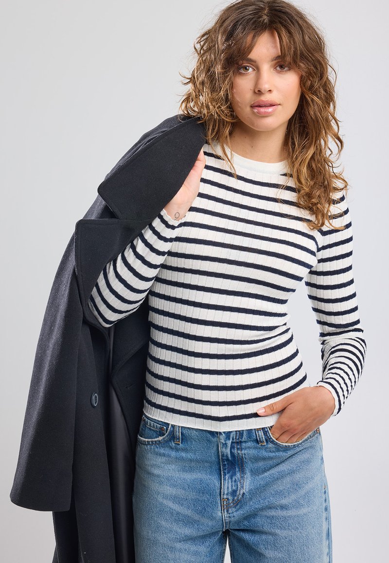 White long-sleeve top with black horizontal stripes, ribbed texture. Paired with light blue high-waisted jeans and a black coat draped over one shoulder.