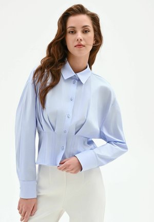 Donna con lunghi capelli castani che indossa una camicia azzurro chiaro abbottonata e pantaloni bianchi, in piedi davanti a uno sfondo bianco uniforme.