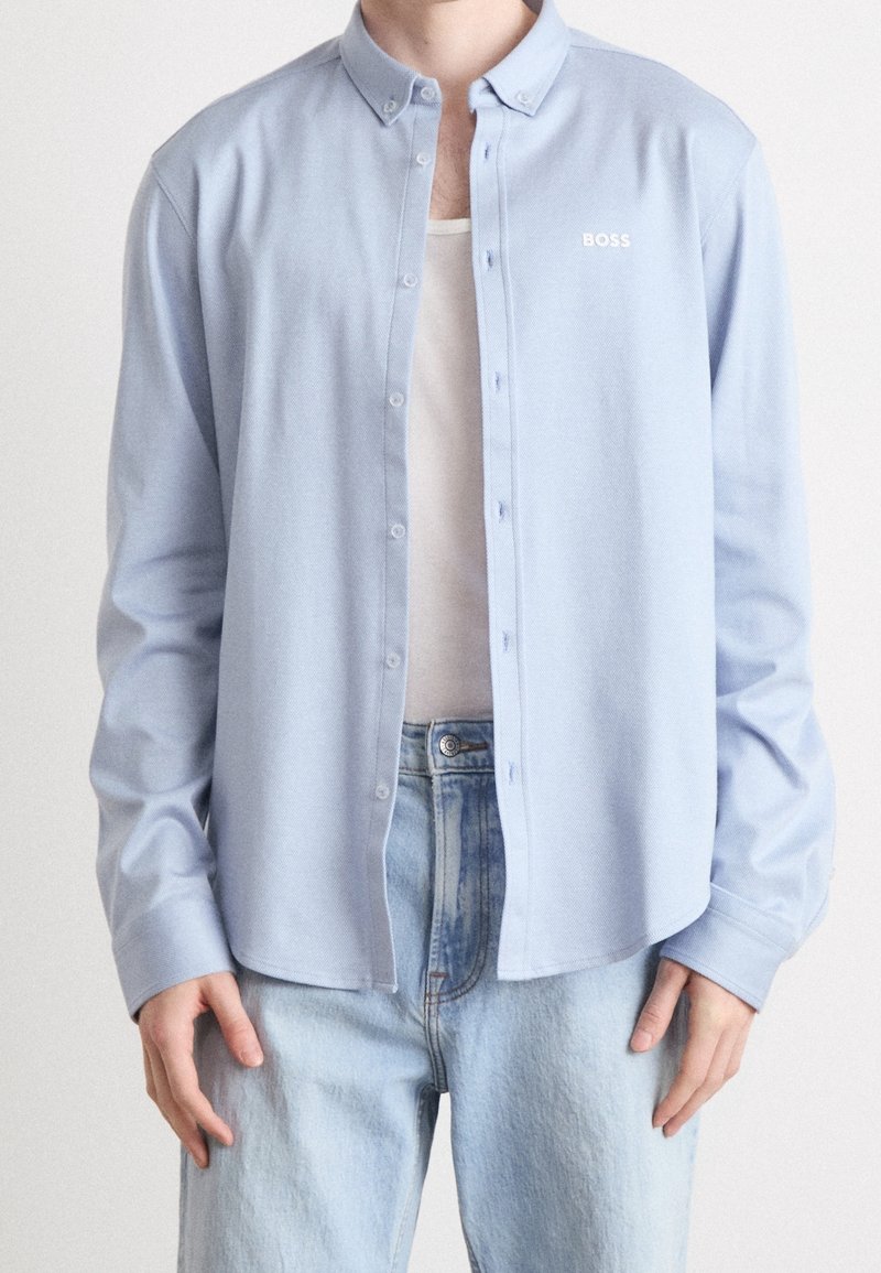 Długa jasnoniebieska koszula na guziki z kołnierzykiem, z małym białym logo, noszona na białym topie i jasnych dżinsach.