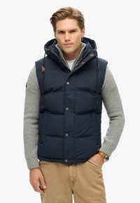 Navy blue puffer vest with a hood, snap buttons, and drawstrings. Worn over a light grey knit sweater. Features visible texture and layers.