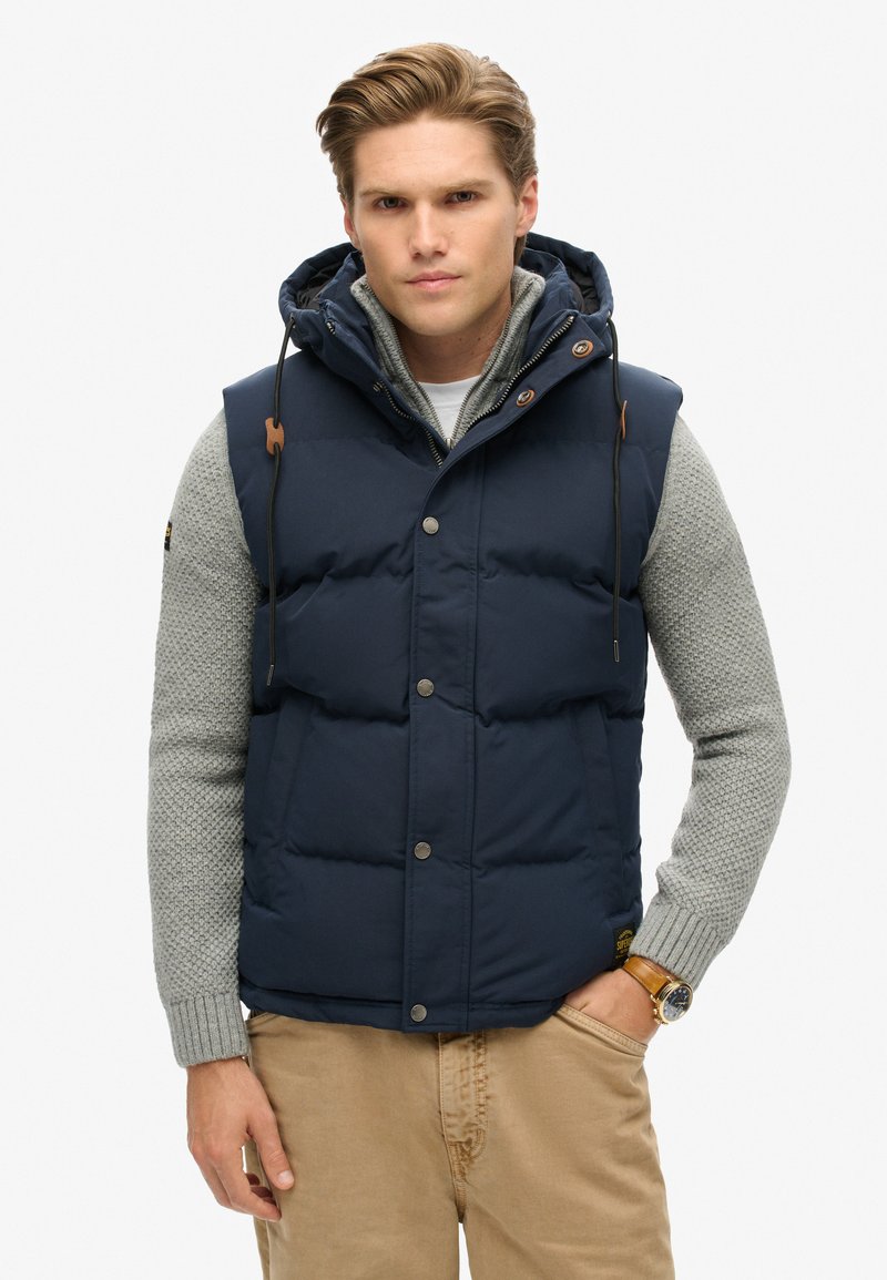 Navy blue puffer vest with a hood, snap buttons, and drawstrings. Worn over a light grey knit sweater. Features visible texture and layers.