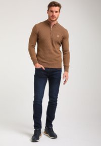 Brauner Strickpullover mit einem halben Reißverschlusskragen, der mit einem strukturierten Rautenmuster versehen ist, kombiniert mit dunkelblauen Jeans und navyfarbigen Turnschuhen.