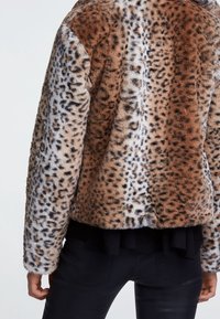 Veste en fausse fourrure avec un imprimé léopard mêlant des couleurs marron et crème. Coupe courte avec une texture douce. Un pantalon noir est visible.