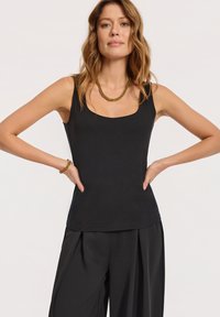 Kobieta o falowanych włosach w bezrękawnikowej czarnej bluzce i szerokich czarnych spodniach, uzupełniona złotym łańcuszkiem na szyi i bransoletką, z rękami na biodrach.