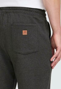 Nærbilde av mørkegrå joggebukser med elastisk midje og baklomme som har et lite brunt logomerke, båret av en person i hvit skjorte.
