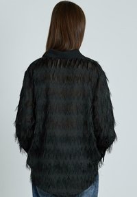 Blusa negra transparente con patrón de zigzag, acabado texturizado y mangas largas. Cuenta con un collar negro y un corte suelto y relajado.