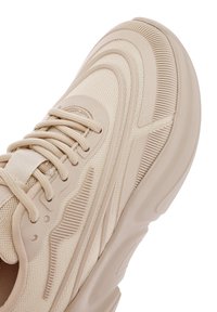 Beige Sportschuh mit Obermaterial aus Mesh, strukturierten Details, Gummisohle und Schnürdesign, das dezente Wellenmuster und ausgeprägte Akzente aufweist.