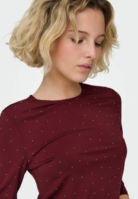 Kobieta o krótkich kręconych blond włosach, ubrana w bordową koszulę w drobne białe kropki, patrząca w dół z neutralnym wyrazem twarzy.